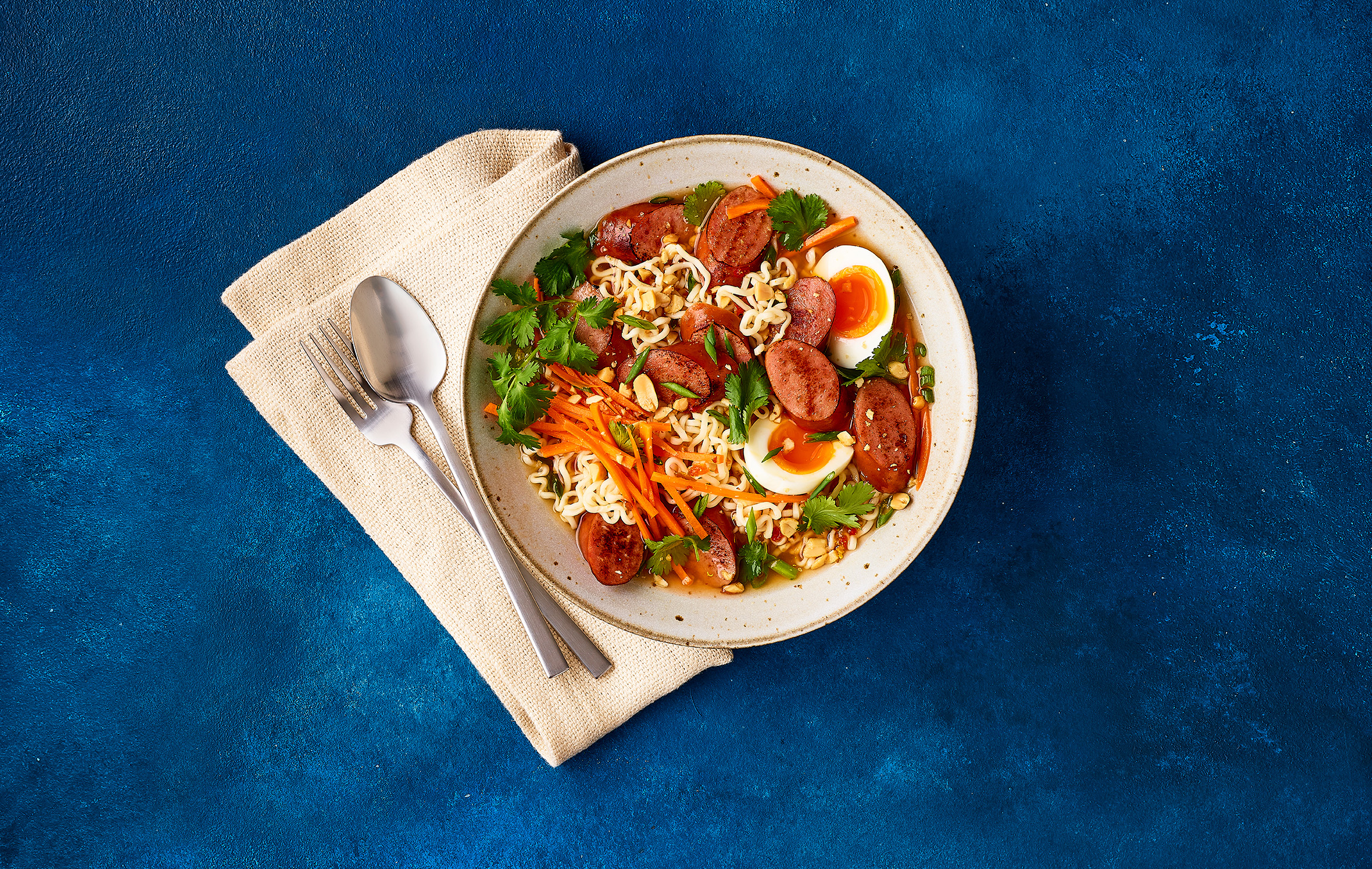 Bowl of ramen with spicy turkey sausage, eggs, carrots, cilantro, and peanuts on a blue background.