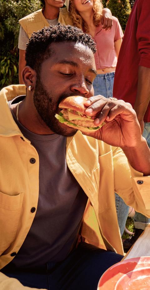 Man eating a turkey burger at an outdoor gathering with friends on a sunny day